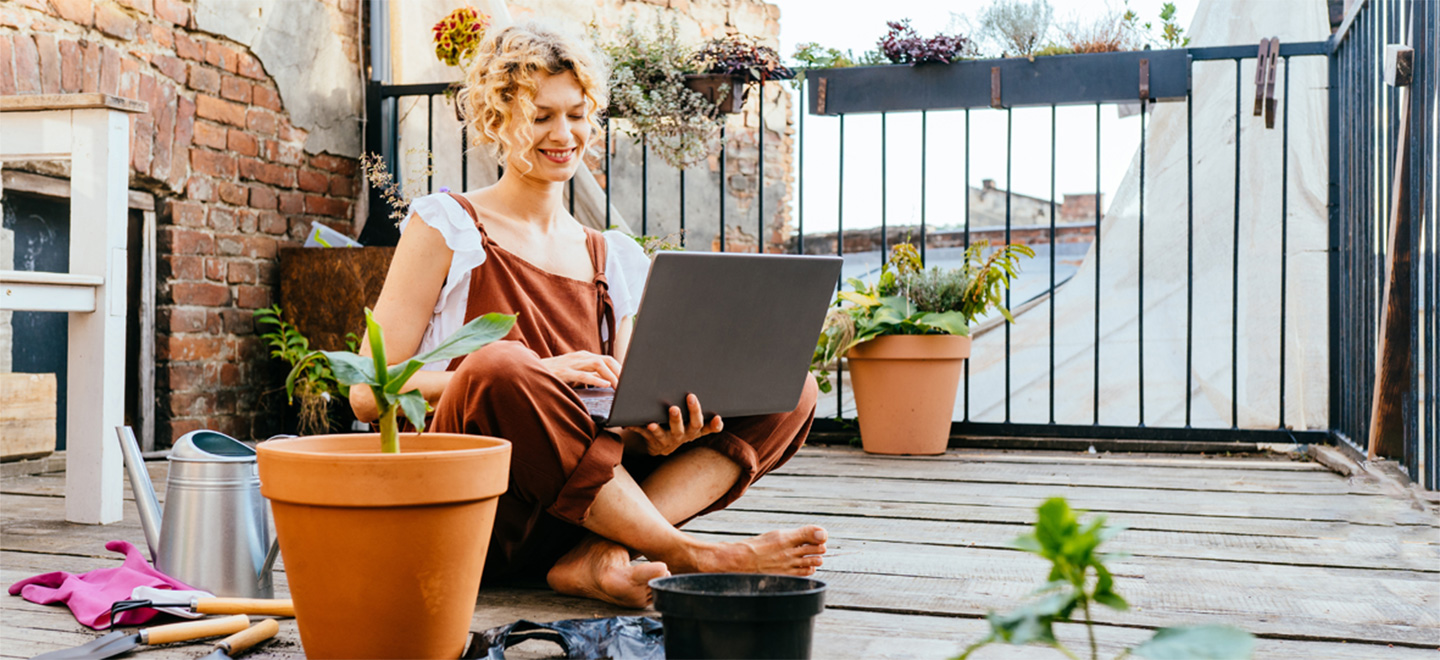 Eine blonde Frau sitzt auf einem Balkon im Schneidersitz und hat einen Laptop in der Hand. Um sie herum stehen Blumentöpfe und Werkzeuge zum Gärtnern.