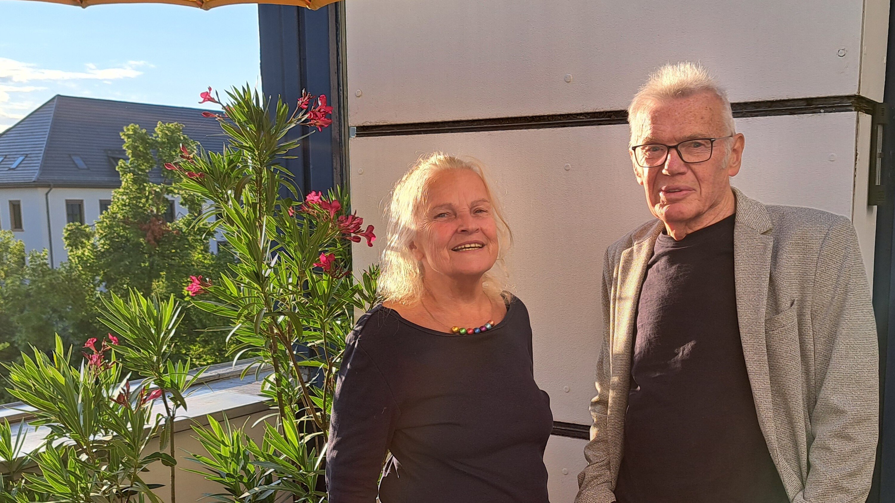 Ein älteres Paar steht auf einem sonnigen Balkon neben blühenden Pflanzen. Beide lächeln in die Kamera, im Vordergrund sind bunte Blumen und ein gelber Sonnenschirm zu sehen.