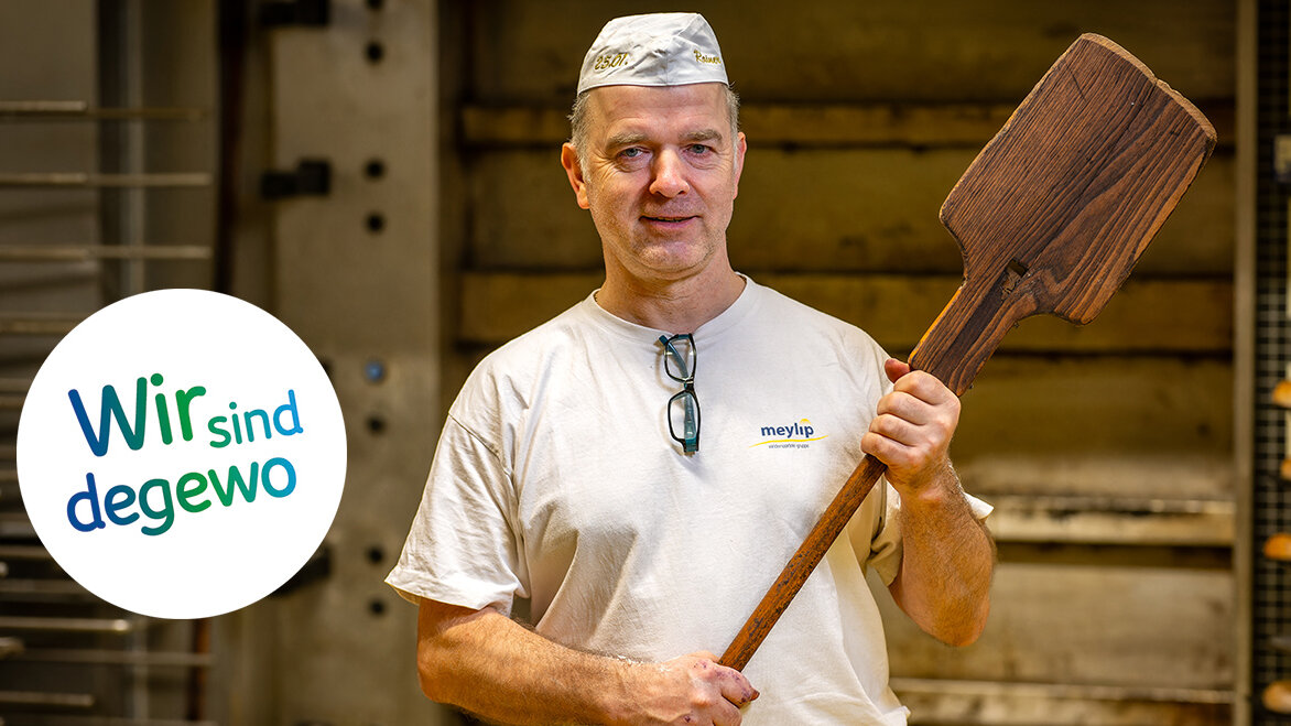 Ein Bäcker steht vor dem Ofen, hält eine große Holzschaufel und trägt Bäckermütze und Arbeitshemd.