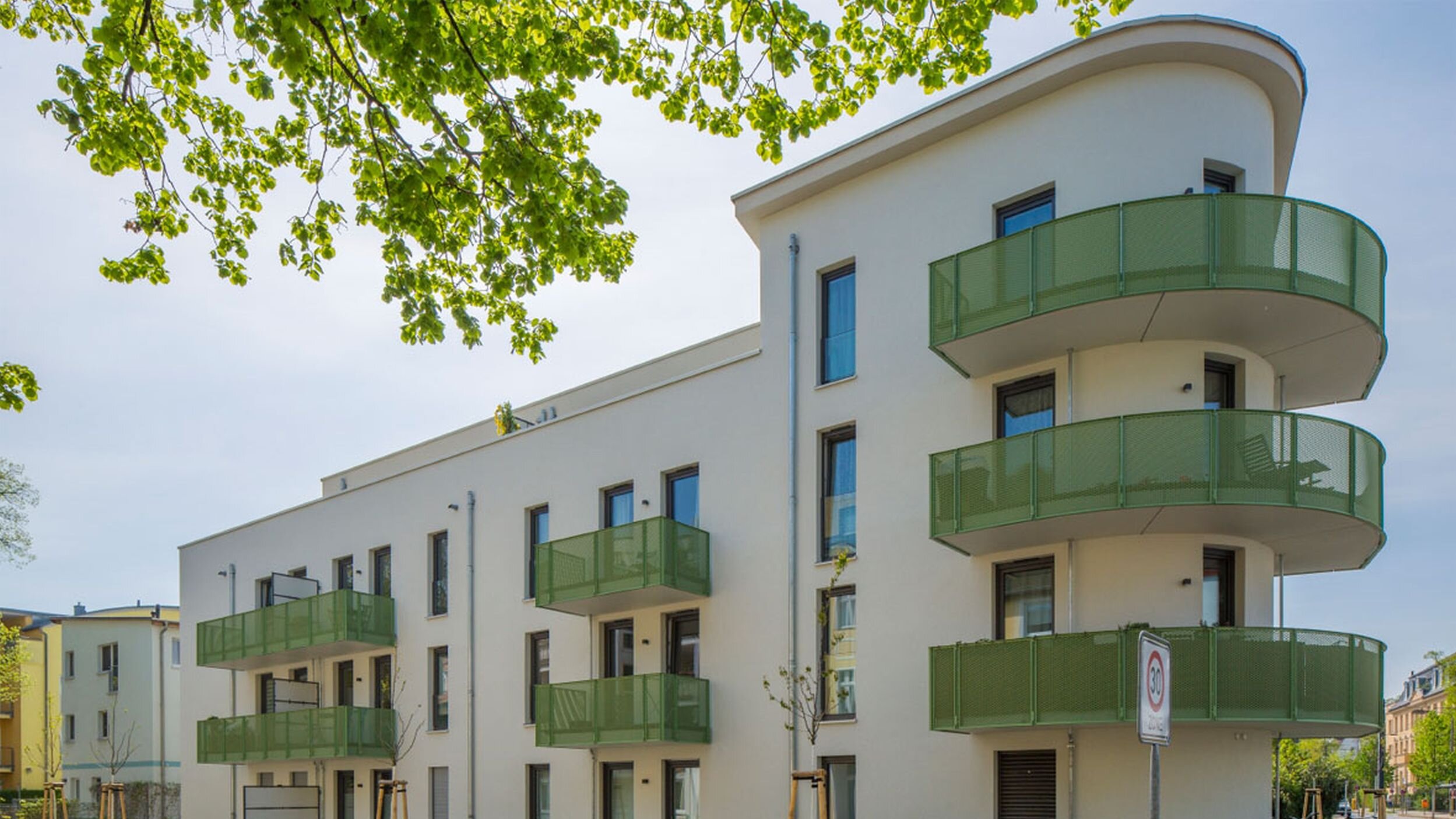 Straßenansicht degewo-Neubau Wassersportallee 13 mit blättrigen Baumzweigen oben im Vordergrund.