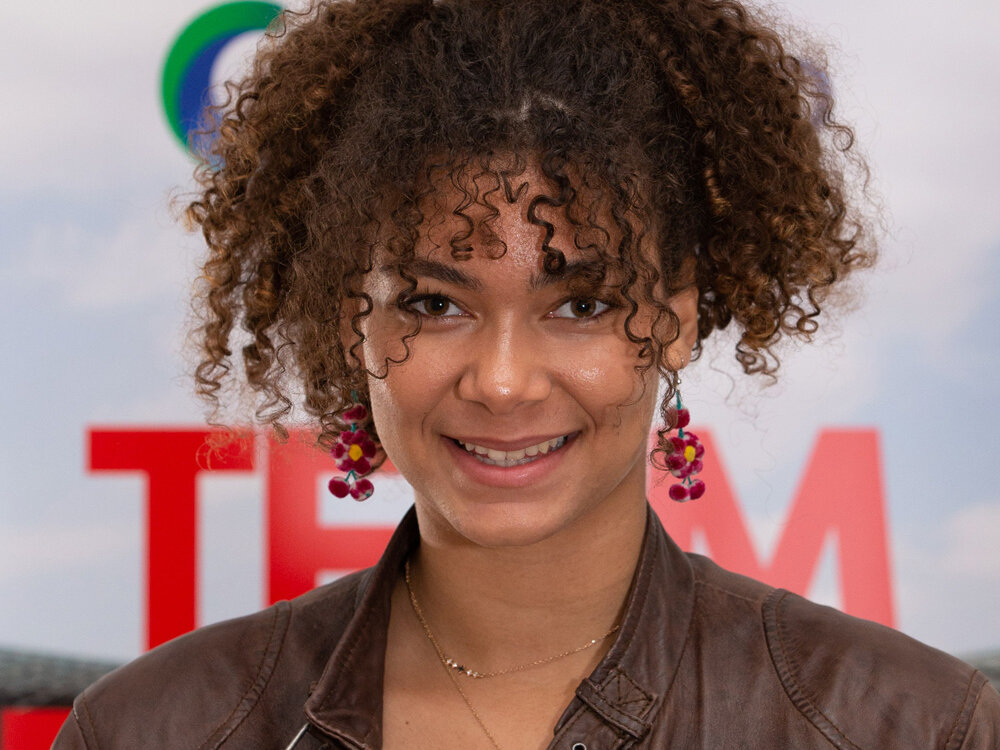Junge Frau mit lockigem Haar und brauner Lederjacke steht vor einer Sponsorenwand und lächelt freundlich.