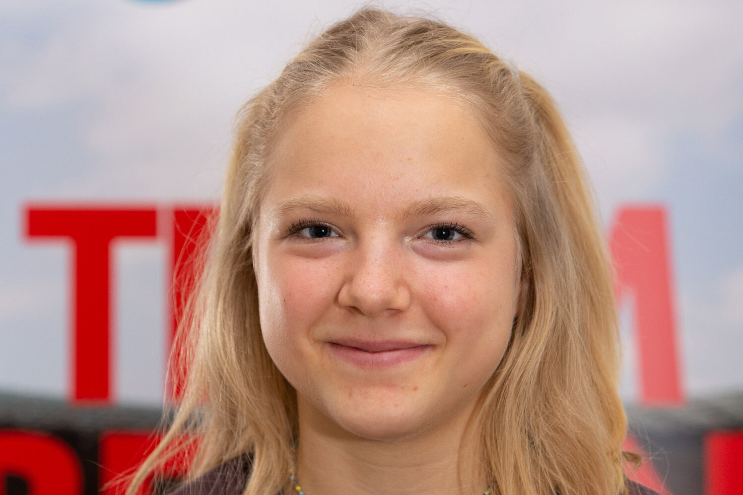 Junge Sportlerin mit blondem Haar steht vor einer Sponsorenwand und lächelt freundlich.