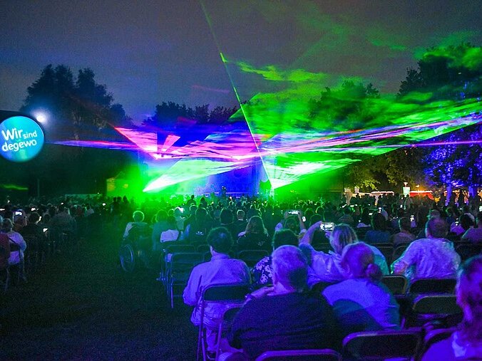 Man sieht eine Lasershow vor großer Bühne. Auf einer großen Rasenfläche davor sitzen oder stehen Menschen und schauen gespannt dabei zu. 