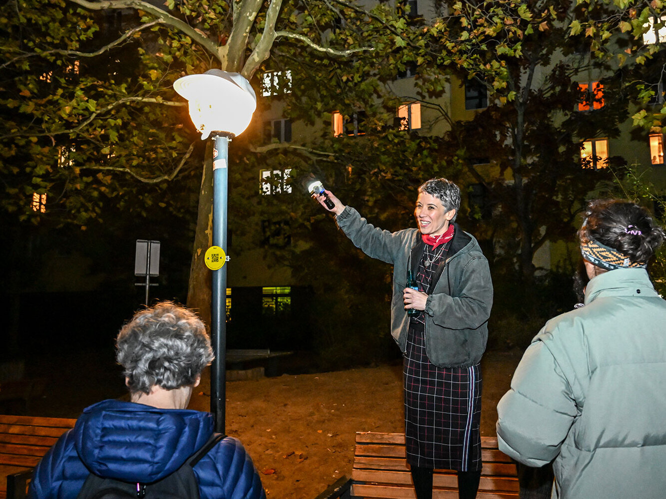 Eine Frau führt eine kleine Gruppe bei Dunkelheit an einer beleuchteten Straßenleuchte vorbei. Die Laterne trägt eine helle Mützen-Verkleidung und die Gruppe schaut interessiert zu.