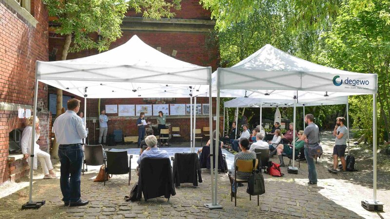 Menschen stehen unter einem Pavillon in einem Hof und besprechen Pläne, die vor ihnen auf dem Boden liegen.