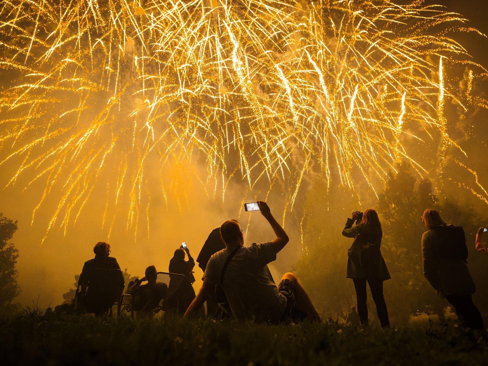 Besucherinnen und Besucher beobachten ein großes Feuerwerk am Nachthimmel. Mehrere Personen fotografieren oder filmen die goldenen Feuerwerkseffekte mit ihren Smartphones.