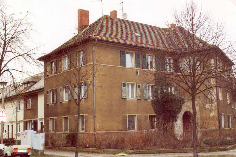An einer Straßenecke steht ein unsaniertes Wohnhaus mit dunkler Fassade, alten Fensterläden und bröckelndem Putz. Davor kahle Pflanzen und ein Bürgersteig.