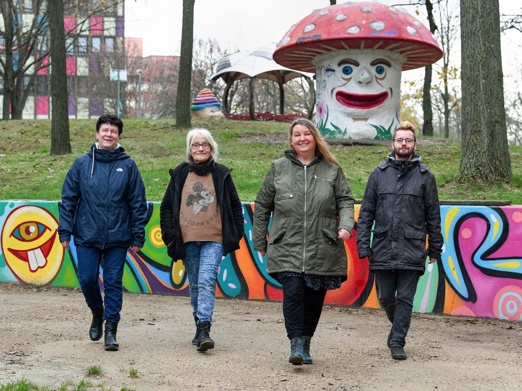 Vier Personen gehen lächelnd durch einen farbenfroh bemalten Park. Im Hintergrund steht eine große Pilzskulptur, sowie einige Spielgeräte.