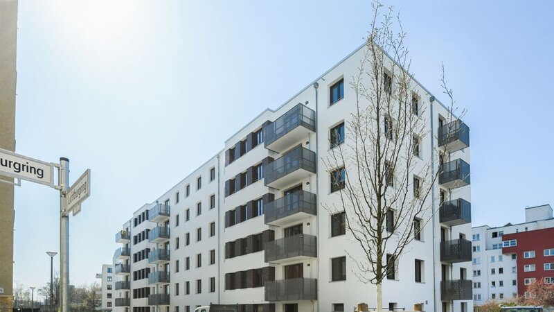 Straßeneckenansicht degewo-Neubau Lichtburgring mit Straßenschild und neu gepflanztem Baum im Vordergrund.