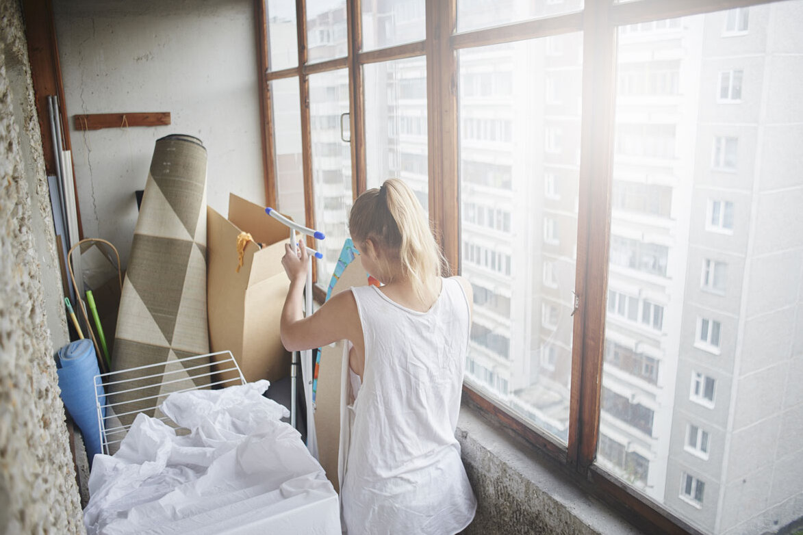 Frau steht in einer Loggia und räumt Gegenstände wie Pappen, Textilien und Haushaltsutensilien zusammen; durch die Fensterfront sind benachbarte Wohngebäude sichtbar.