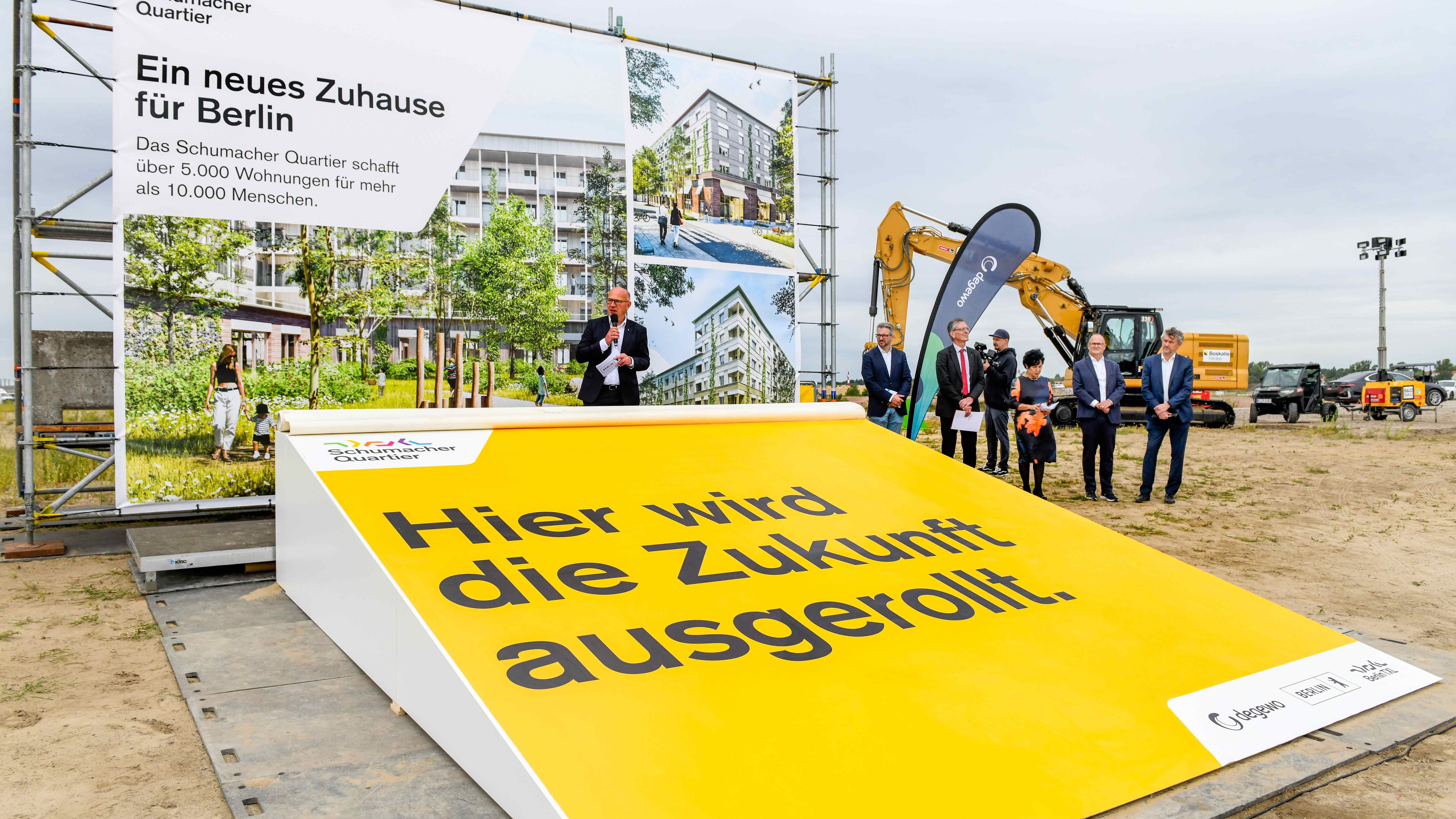 Blick auf ein großes Plakat auf einer Baustelle. Auf der Plakat steht folgender Text: Ein neues Zuhause für Berlin. Dazu das Logo des Schumacher Quartiers und Fotos, wie das künftige Quartier aussehen soll. Neben dem Plakat ein degewo-Aufsteller und mehrere Personen.