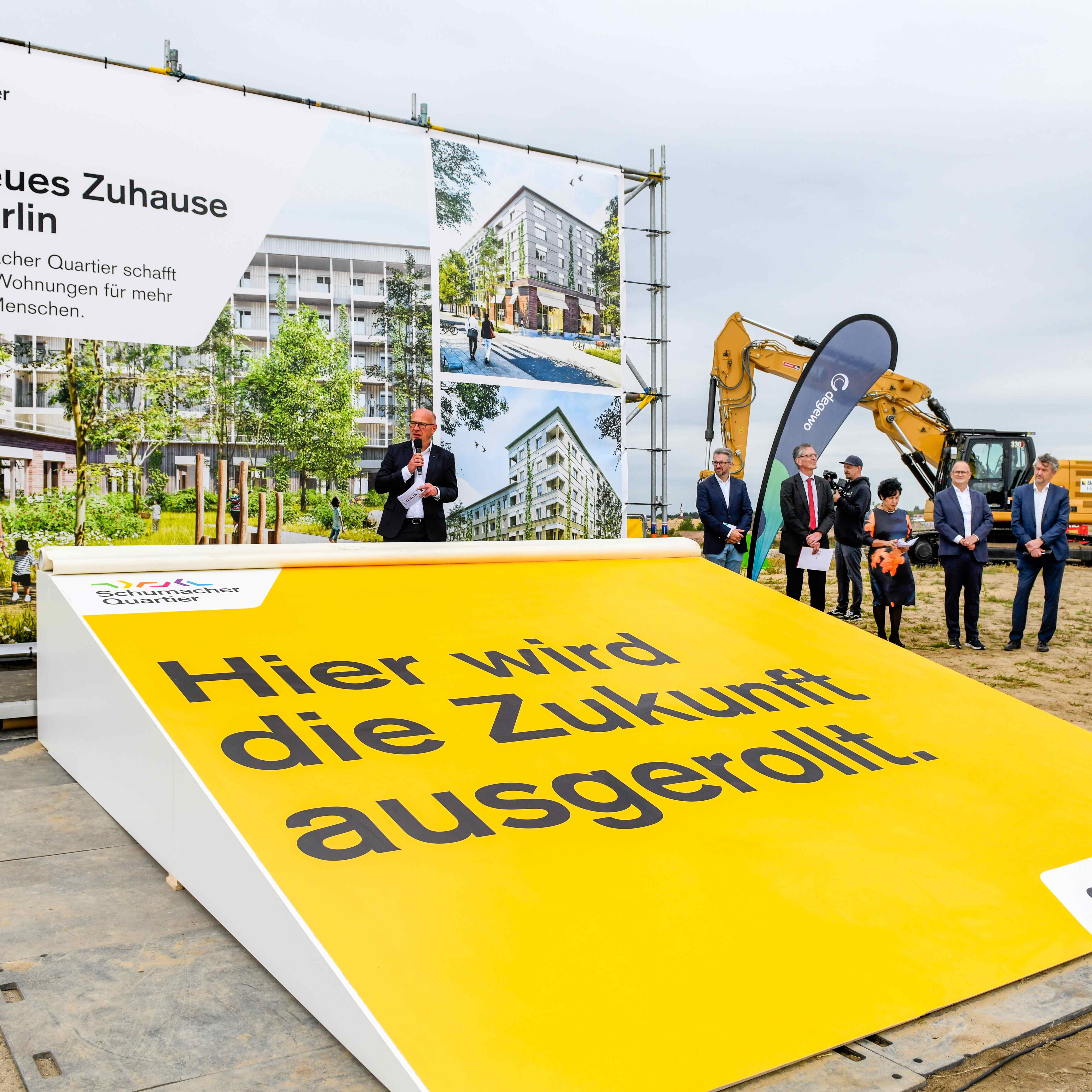 Blick auf ein großes Plakat auf einer Baustelle. Auf der Plakat steht folgender Text: Ein neues Zuhause für Berlin. Dazu das Logo des Schumacher Quartiers und Fotos, wie das künftige Quartier aussehen soll. Neben dem Plakat ein degewo-Aufsteller und mehrere Personen.