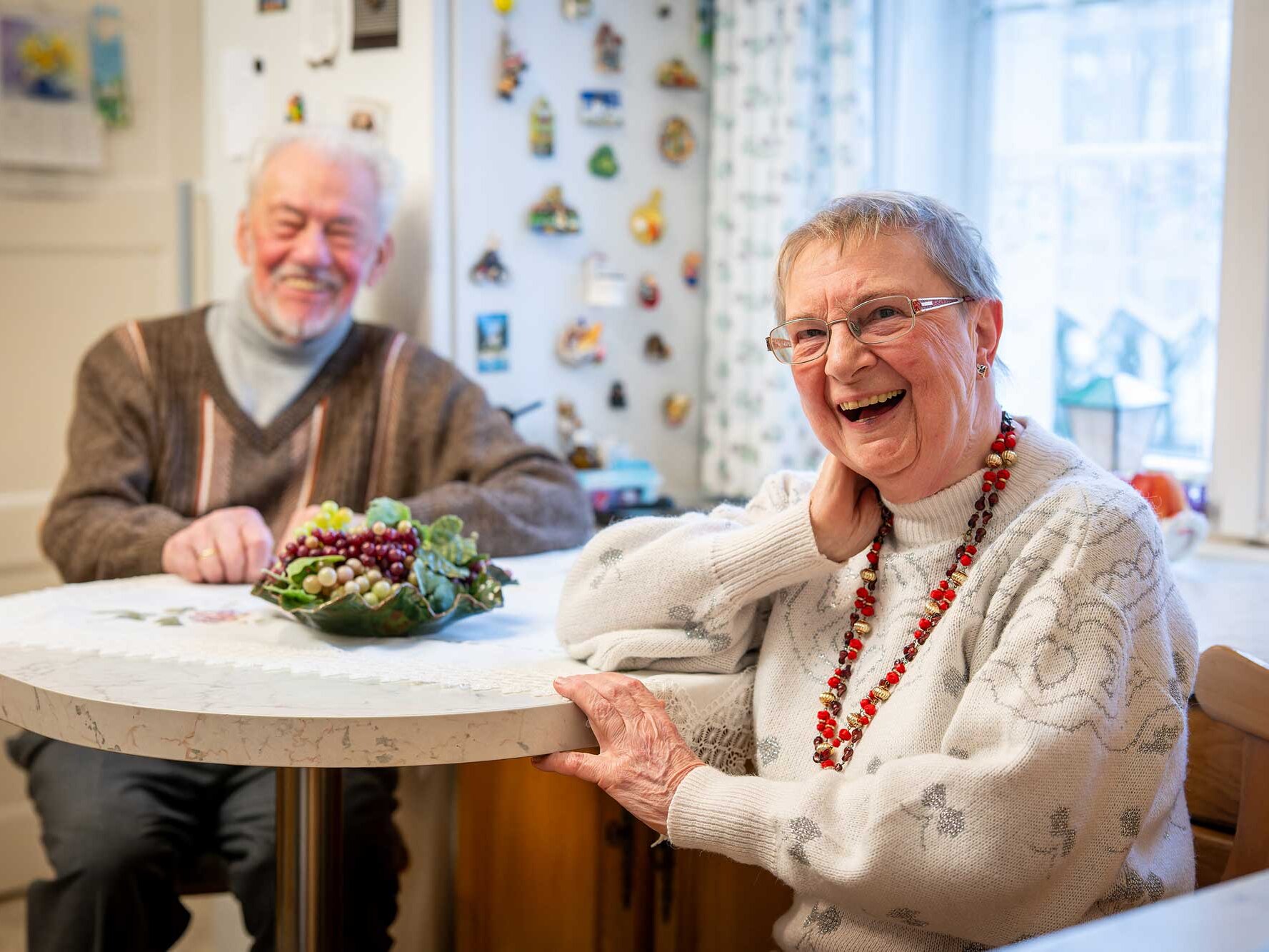 Ein älteres Paar sitzt lachend in einer gemütlichen Küche am Tisch.