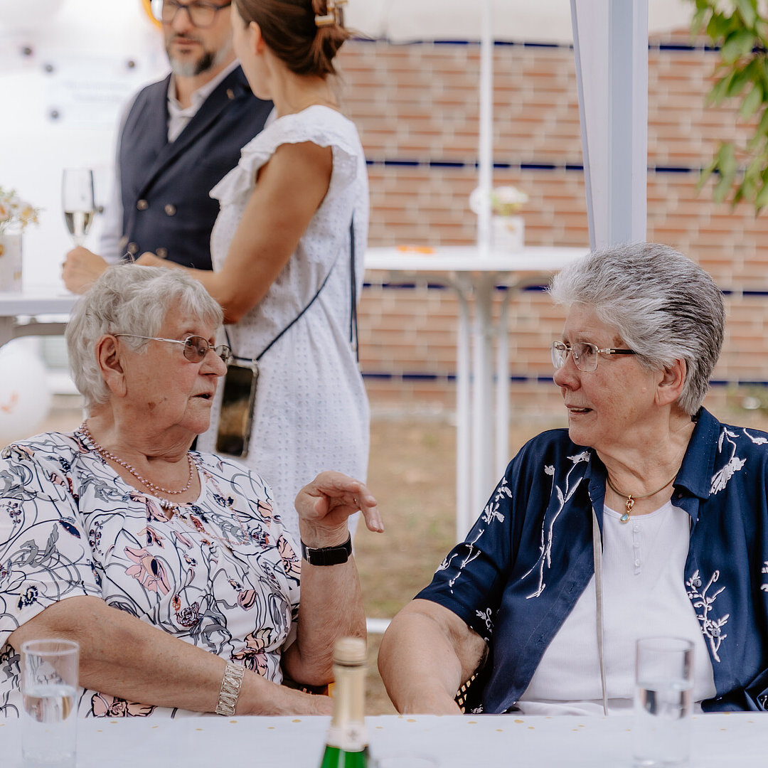 Zwei Seniorinnen sitzen an einem Tisch nebeneinander und unterhalten sich.