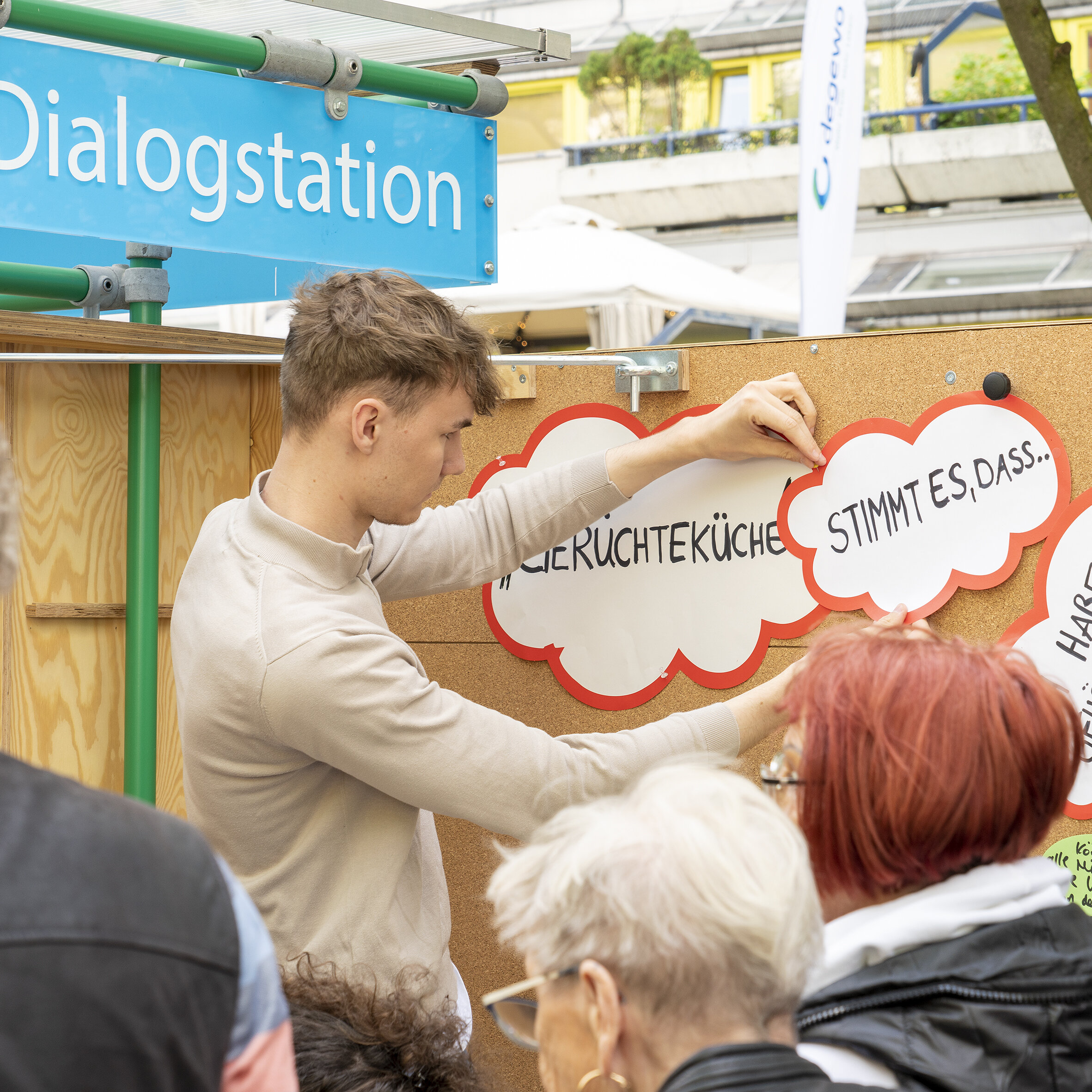 Ein junger Mann steht vor einer Korkwand und pinnt Zettel an die Wand. Auf den Zetteln stehen Teilsätze wie. Stimmt es, dass …, Gerüchteküche, Habe gehört, dass … Vor der Wand stehen mehrere Personen. 
