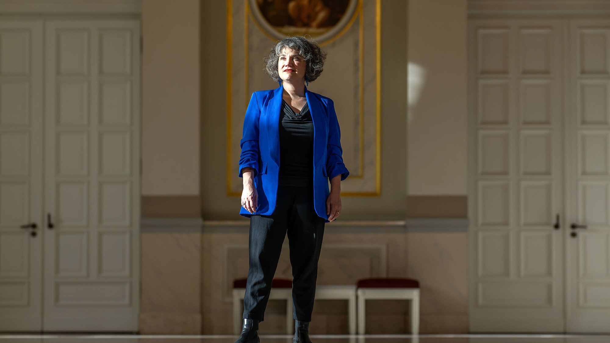 Eine Frau steht in einem großen historischen Saal und trägt ein blaues Sakko. Sie blickt leicht nach oben und wird vom Sonnenlicht angestrahlt.