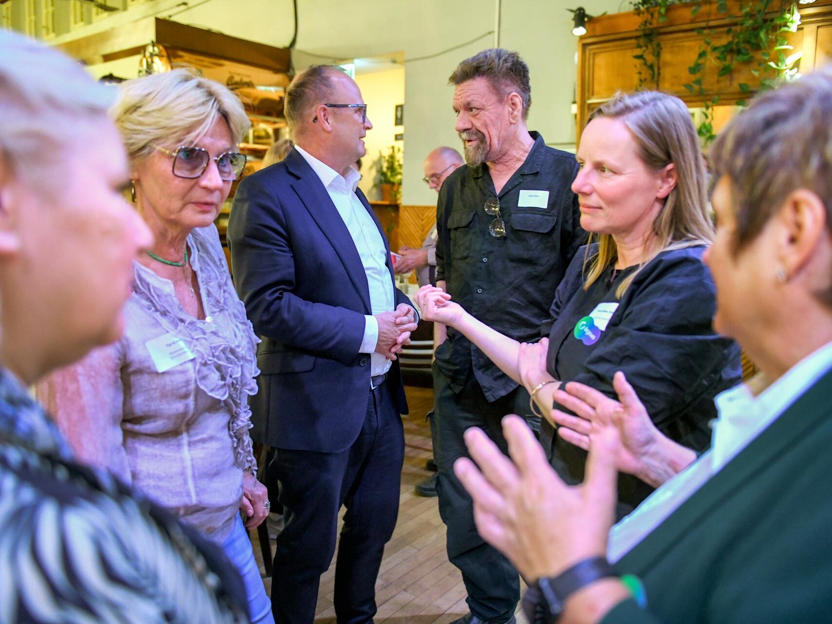 Mehrere Teilnehmende einer Veranstaltung stehen in einem Innenraum zusammen und tauschen sich in einer Gesprächsrunde aus. Im Hintergrund sind weitere Gäste sowie die warm gestaltete Einrichtung des Veranstaltungsortes zu sehen.