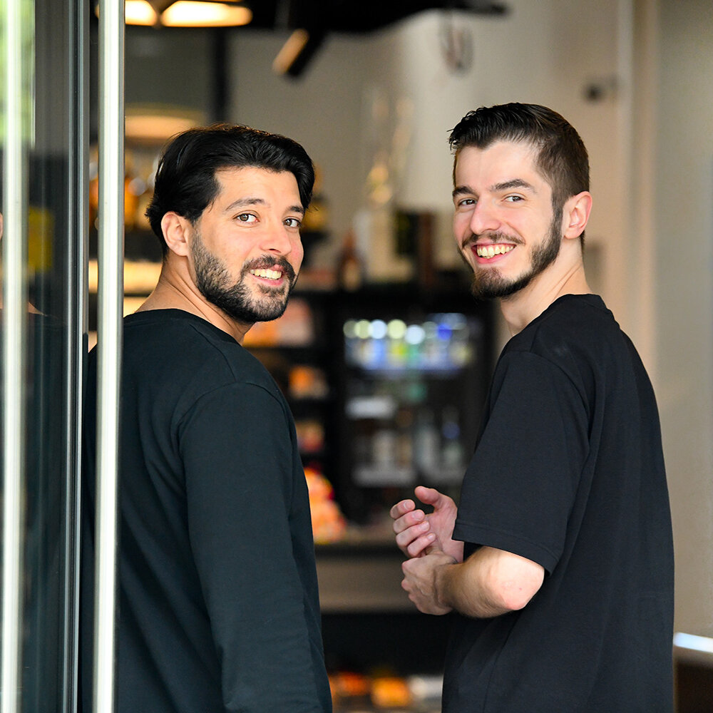 Zwei Männer stehen im Eingang ihres Kiezshops und blicken zurück in die Kamera. Sie lächeln.
