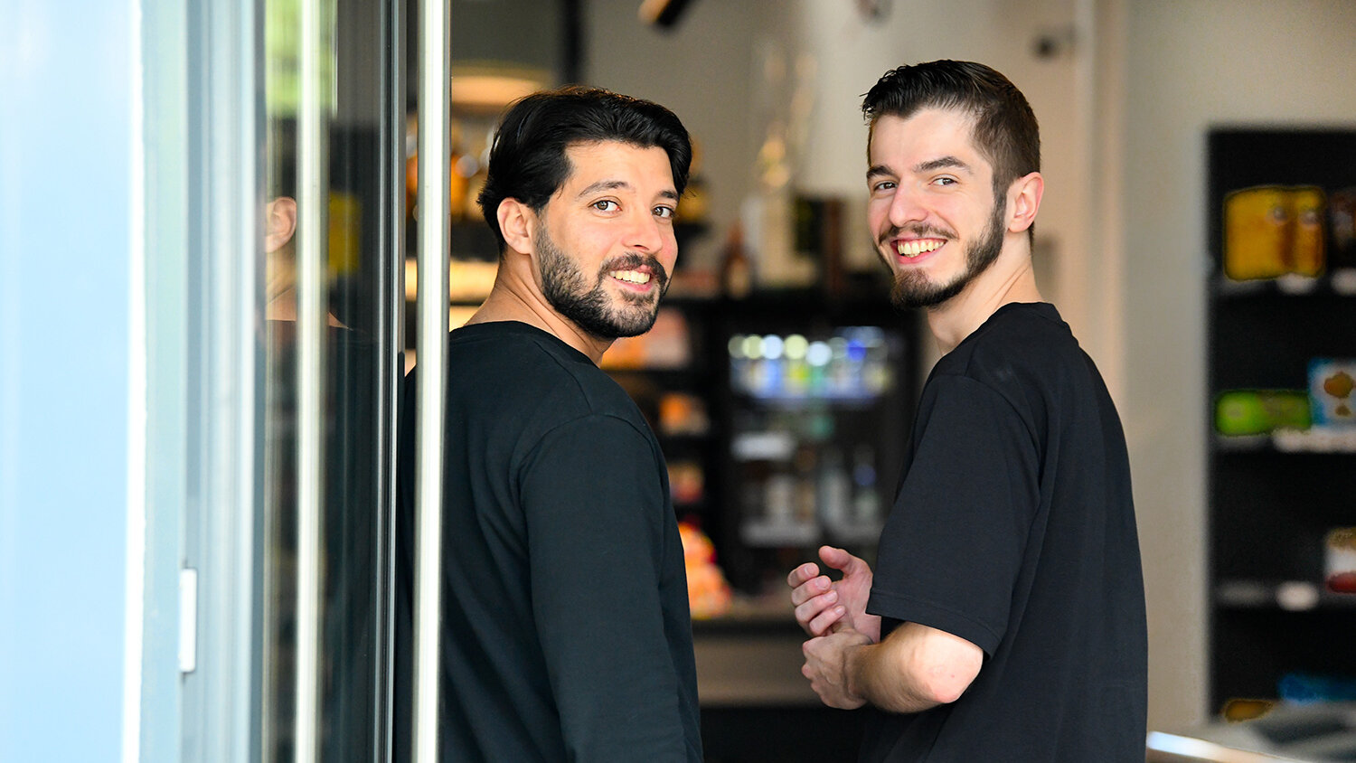 Zwei Männer stehen im Eingang ihres Kiezshops und blicken zurück in die Kamera. Sie lächeln.