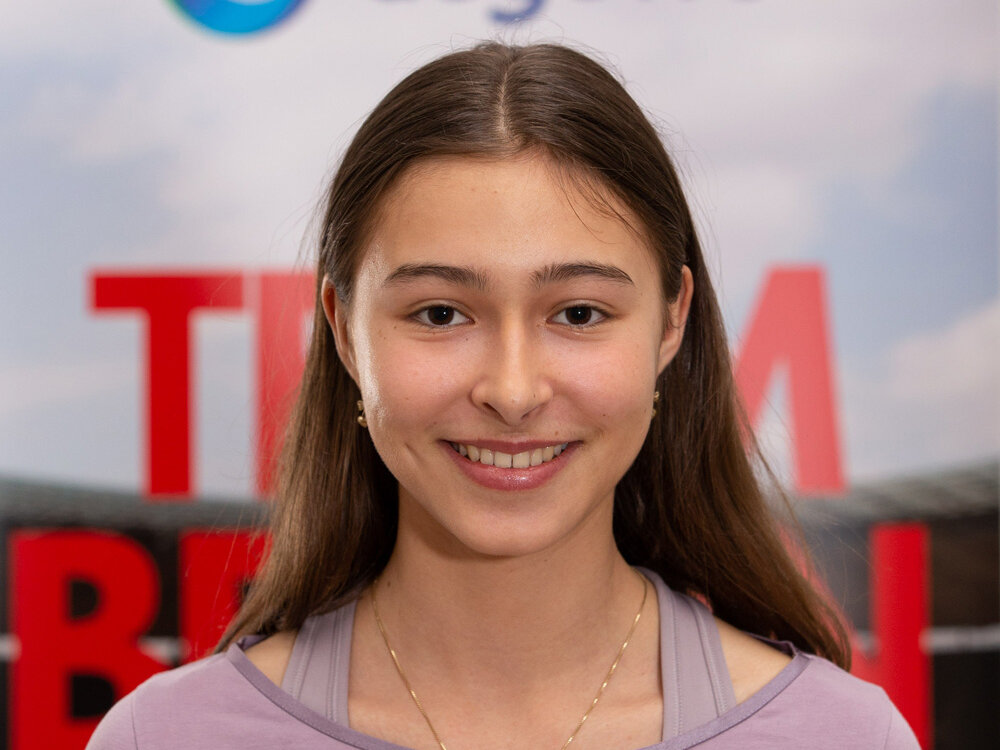 Junge Sportlerin trägt ein lila Oberteil und lächelt vor einer Sponsorenwand mit LOTTO Berlin und degewo Logos.