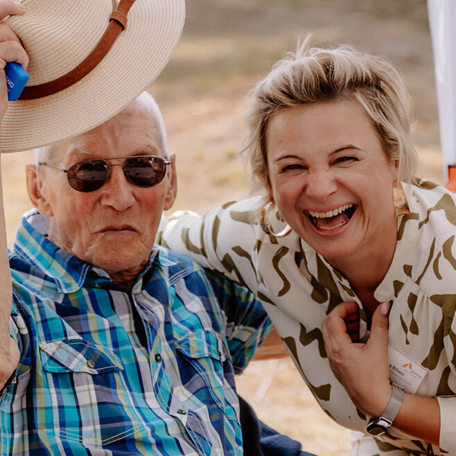 Eine ältere Person mit Sonnenhut und Sonnenbrille sitzt lächelnd neben einer jüngeren Frau von SOPHIA, die herzlich lacht. Beide wirken fröhlich und entspannt in einer sommerlichen Außenumgebung. Im Hintergrund sitzt eine weitere ältere Frau.