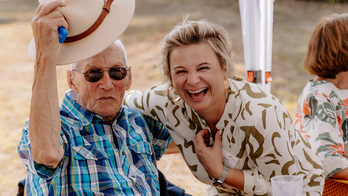 Eine ältere Person mit Sonnenhut und Sonnenbrille sitzt lächelnd neben einer jüngeren Frau von SOPHIA, die herzlich lacht. Beide wirken fröhlich und entspannt in einer sommerlichen Außenumgebung. Im Hintergrund sitzt eine weitere ältere Frau.