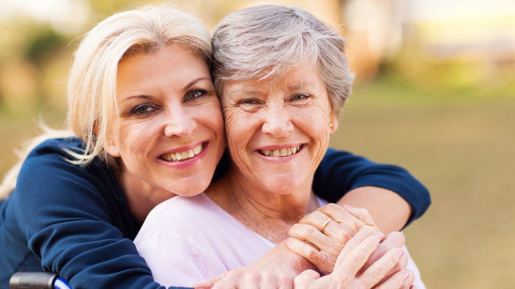Eine Frau umarmt von hinten eine im Rollstuhl sitzende ältere Frau.