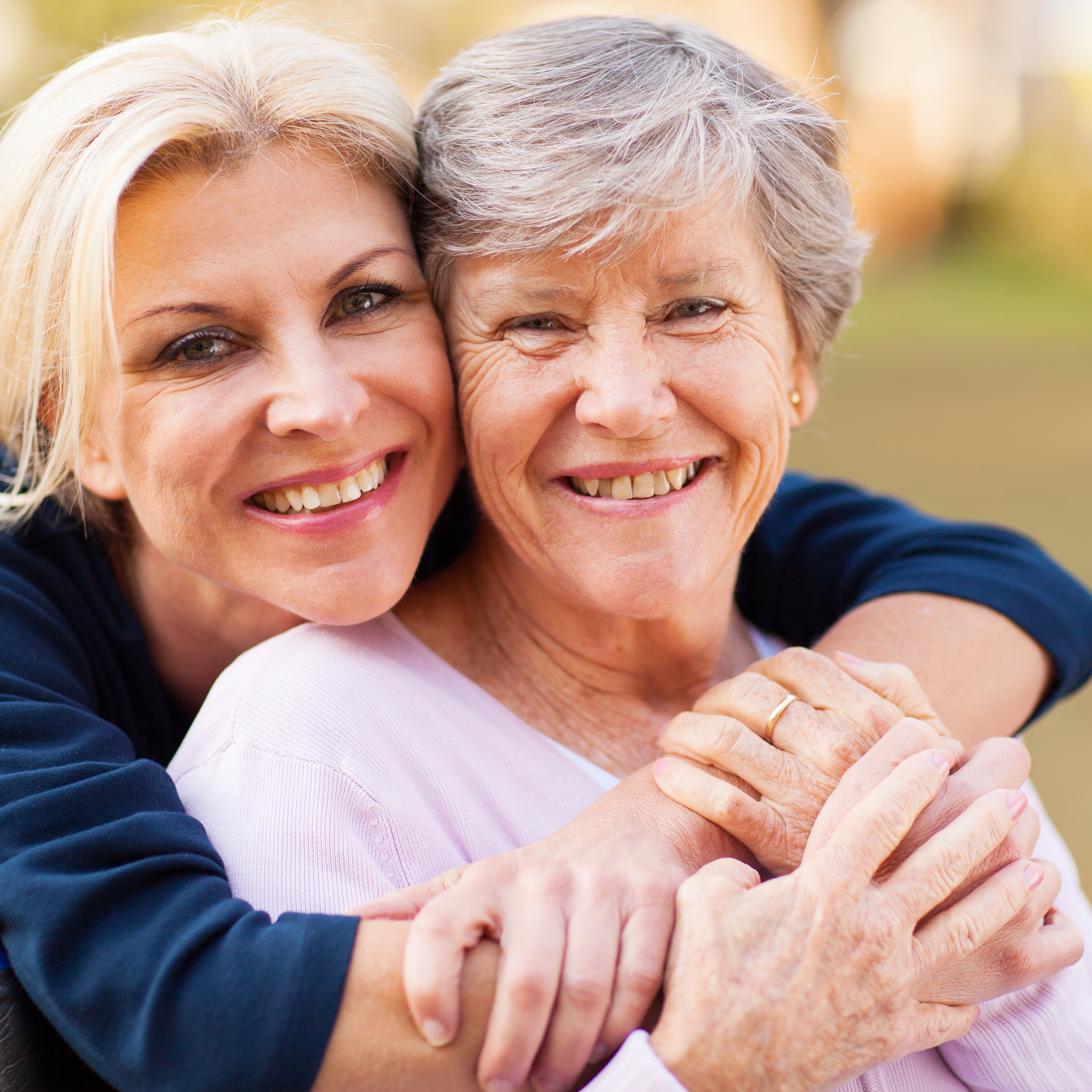 Eine Frau umarmt von hinten eine im Rollstuhl sitzende ältere Frau.