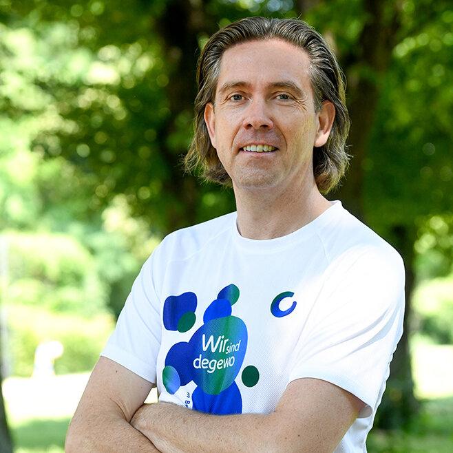 Ein Mann steht in einem Park neben einem Baum. Er trägt ein Sport T-Shirt, seine Arme sind verschränkt.