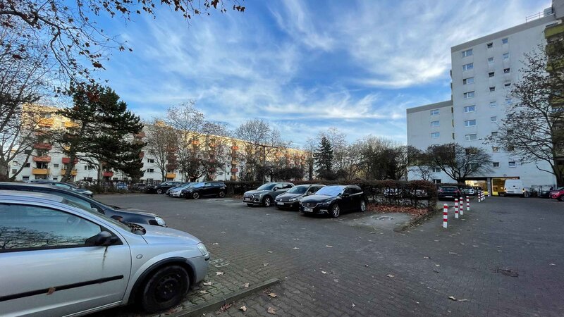Bestehende Parkplatzfläche in einer Wohnsiedlung mit geparkten Fahrzeugen und umliegenden Wohngebäuden. Die Fläche ist größtenteils versiegelt und von Bäumen sowie Grünbereichen eingerahmt.