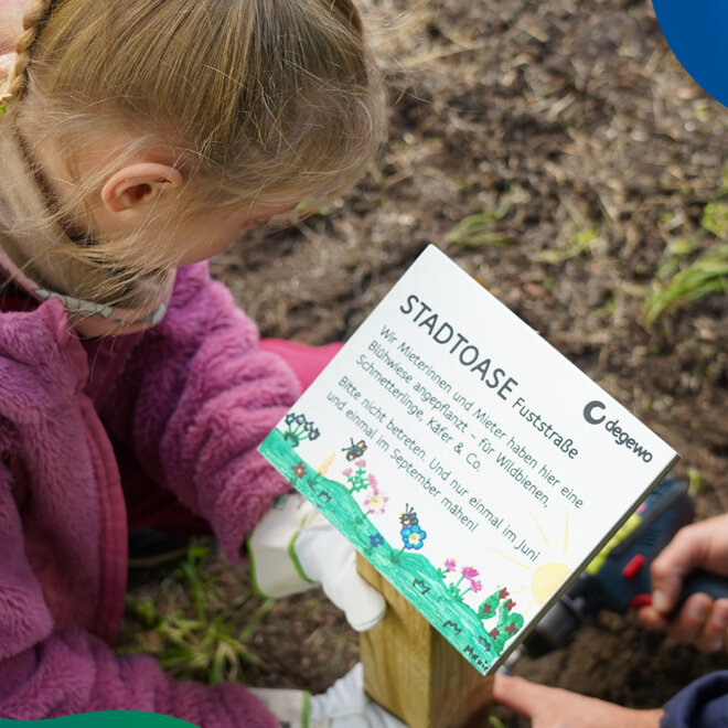 Ein Kind in pinker Jacke befestigt gemeinsam mit einer erwachsenen Person ein Schild im Boden, auf dem "Stadtoase" steht.