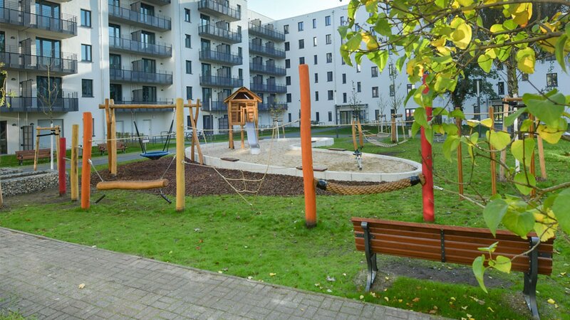 Außenansicht degewo-Neubau Mahlower Straße 2 bis 4 A mit Weg, Sitzbank, Grünfläche, Spielplatz und beblätterten Baumzweigen rechts im Vordergrund.