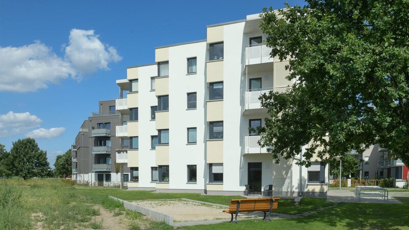 Außenansicht degewo-Neubau Wilhelm-Hoff-Straße 24 und 26 mit Grünfläche, Sandkasten, Sitzbank und Baum rechts im Vordergrund.