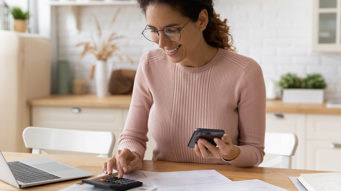 Eine Frau sitzt an einem Küchentisch, hält ein Smartphone in der Hand und tippt gleichzeitig auf einem Taschenrechner. Vor ihr liegen Unterlagen und ein Laptop, während sie lächelnd arbeitet.