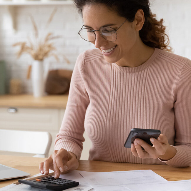 Eine Frau sitzt an einem Küchentisch, hält ein Smartphone in der Hand und tippt gleichzeitig auf einem Taschenrechner. Vor ihr liegen Unterlagen und ein Laptop, während sie lächelnd arbeitet.