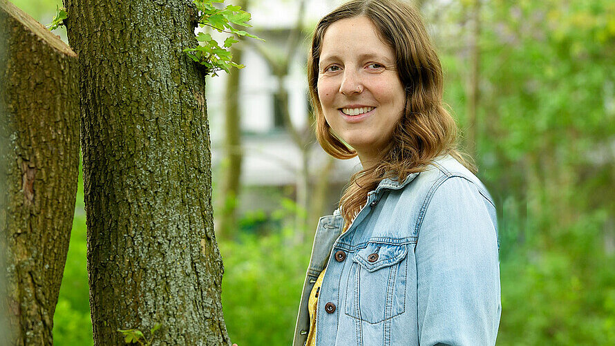 Eine Frau stützt sich mit den Händen an einen Baum ab und lächelt in die Kamera. 
