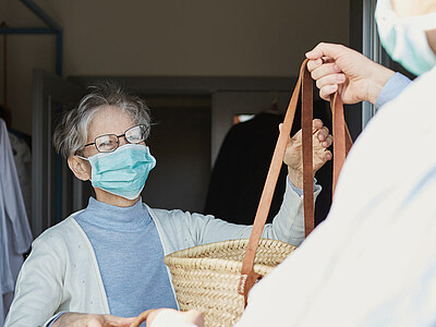 Das Bild zeigt, wie ein Mann einer älteren Frau Einkäufe in Tragekörben übergibt. Beide tragen zum Schutz vor Corona einen Mund-Nase-Schutz.