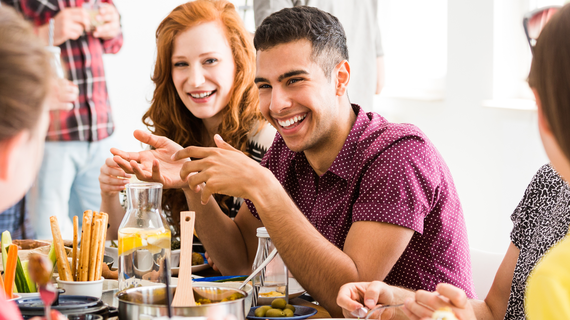 Gruppe junger Menschen sitzt gemeinsam an einem Tisch und unterhält sich gut gelaunt beim Essen.