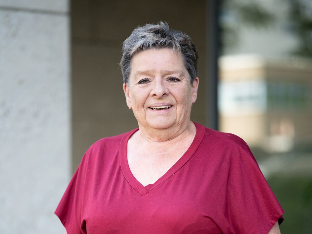 Ältere Frau mit kurzem grauem Haar trägt ein bordeauxfarbenes Oberteil und steht im Freien vor einem Gebäude; sie blickt offen in die Kamera, der Hintergrund ist unscharf.