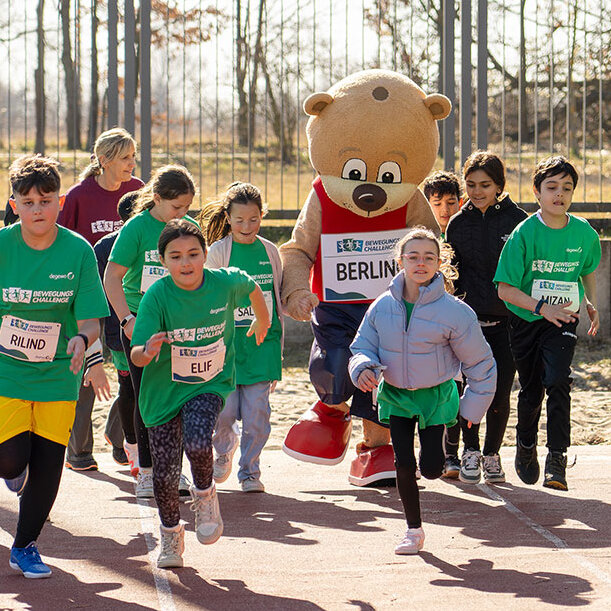 Eine Gruppe Kinder rennt auf einem Sportplatz mit dem Maskottchen Berlino.
