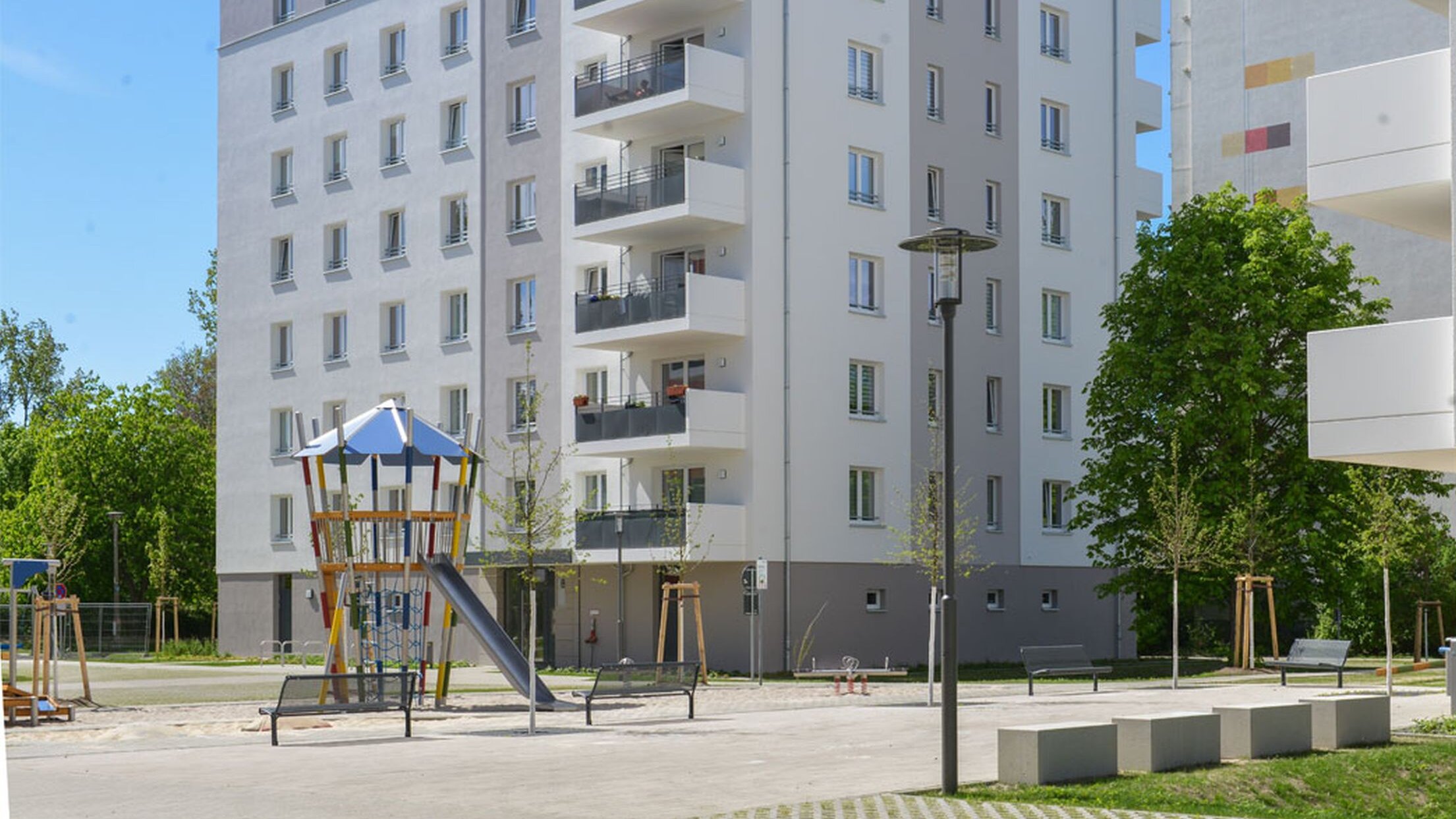 Außenansicht degewo-Neubau Eugen-Roth-Weg 4 bis 10 mit Spielplatz und Straßenlaterne neben Sitzgelegenheiten im Vordergrund.