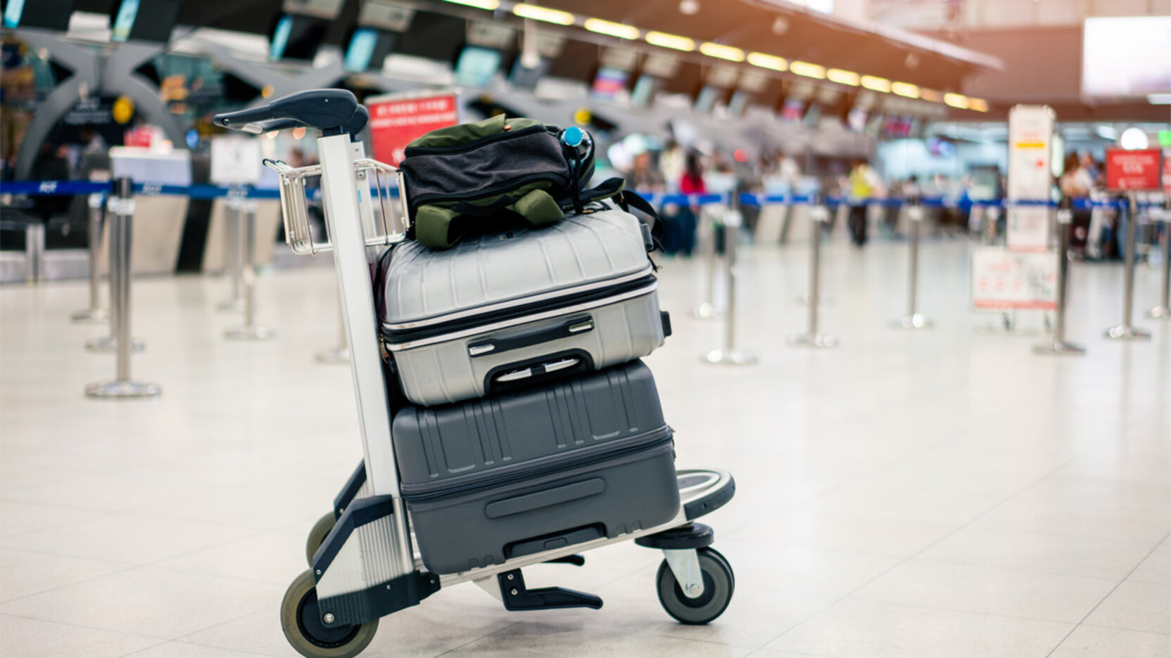 Gepäckwagen mit mehreren Koffern steht in einer Flughafenhalle vor Check-in-Schaltern; im Hintergrund befinden sich Absperrbänder und wartende Reisende.
