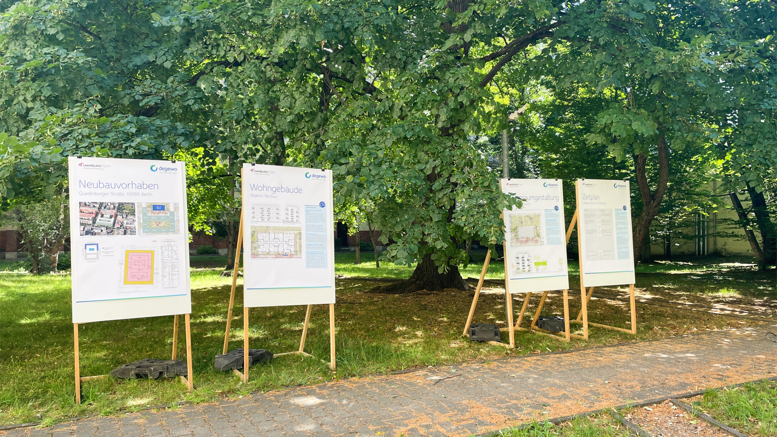 Mehrere Informationsplakate von degewo zum Neubauvorhaben in der Quedlinburger Straße stehen auf Holzstaffeleien im Grünen und informieren Anwohnende über Planung, Wohngebäude, Freiraumgestaltung und Zeitplan.