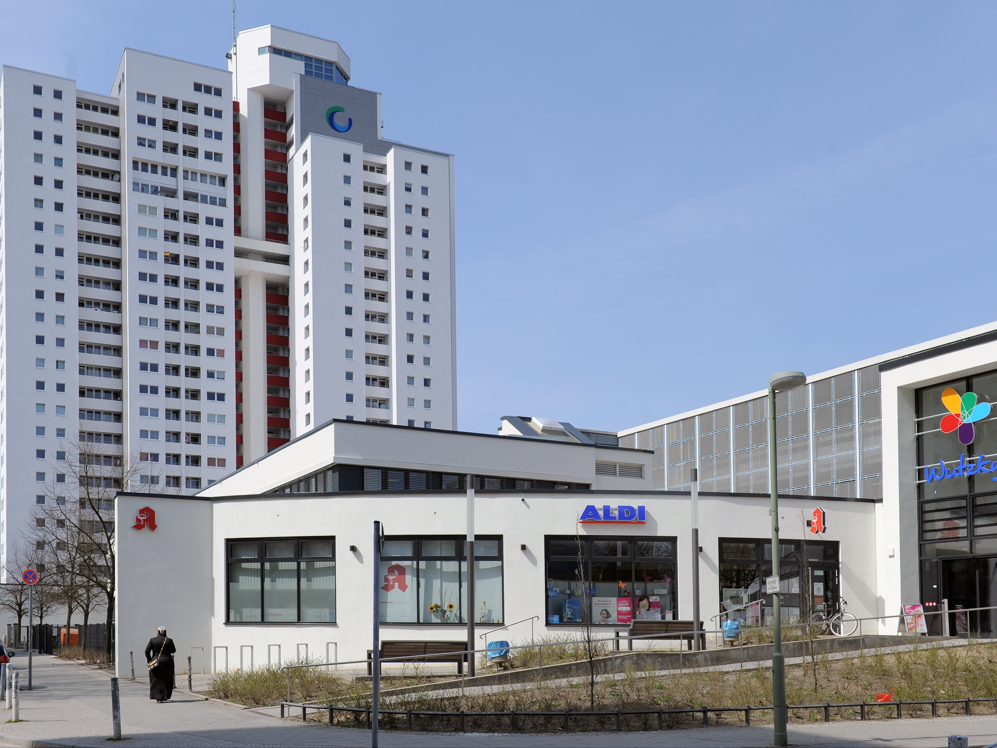 Ein hohes Wohngebäude der Gropiusstadt mit angrenzendem Einkaufszentrum, unter anderem Aldi und eine Apotheke. Im Hintergrund ist ein klarer, blauer Himmel.