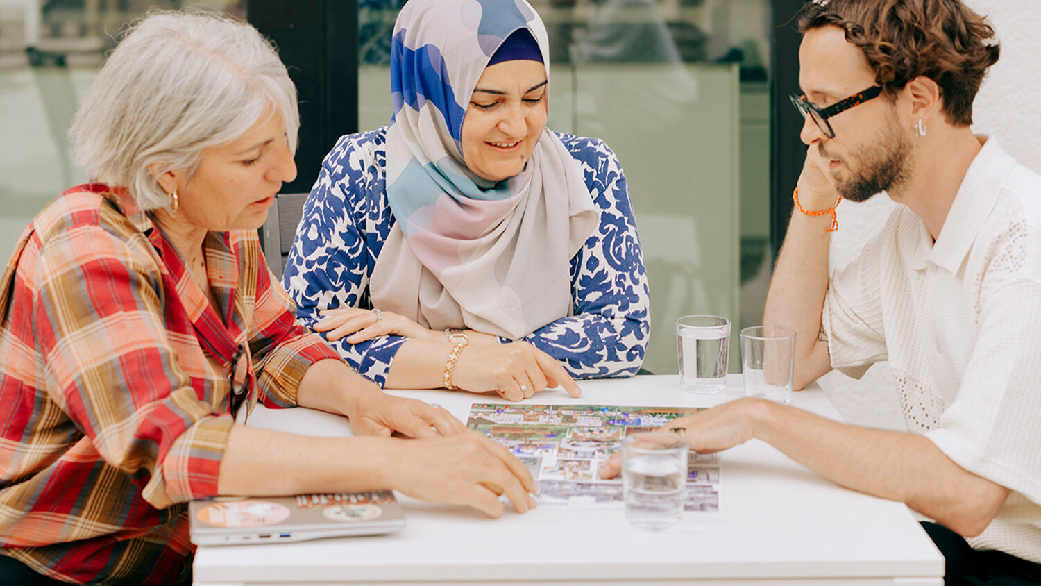Drei Personen sitzen an einem Tisch und beschäftigen sich gemeinsam mit Bildkarten. Die Szene zeigt ein freundliches Gespräch in einer offenen und einladenden Atmosphäre.