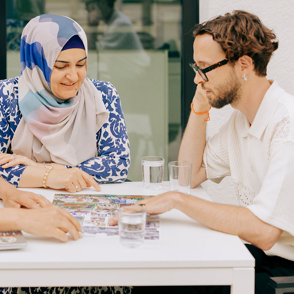 Drei Personen sitzen an einem Tisch und beschäftigen sich gemeinsam mit Bildkarten. Die Szene zeigt ein freundliches Gespräch in einer offenen und einladenden Atmosphäre.