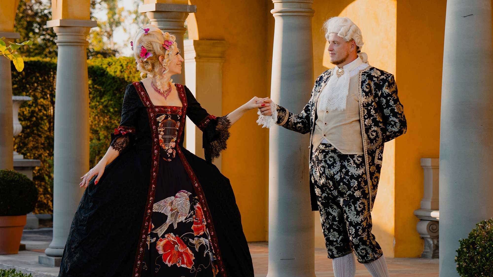 Zwei Personen tragen aufwendige barocke Kostüme und posieren vor Säulen in einer Gartenanlage. Das Kleidungsdesign zeigt florale und vogelartige Motive.