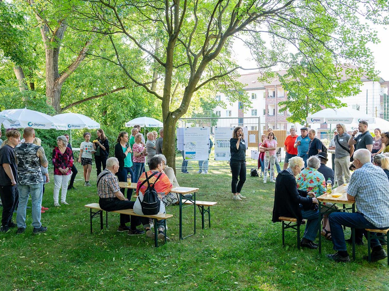Zahlreiche Anwohnende haben sich unter Bäumen versammelt und hören einer Rednerin zu. Einige sitzen an Bierzeltgarnituren, andere stehen, im Hintergrund sind Projektpläne ausgehängt.