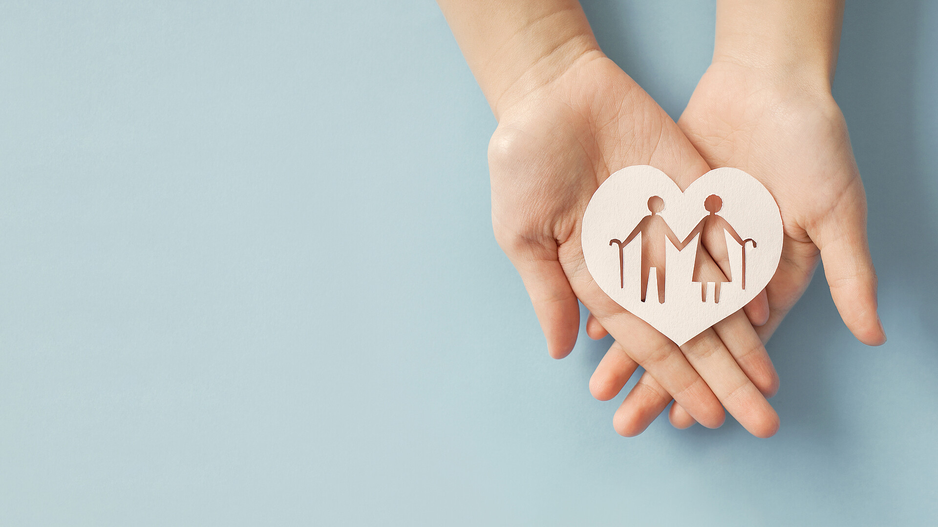 Zwei Hände halten ein Herz aus Pappe in der Hand. Aus dem Herz wurden zwei Personen mit Gehstöcken ausgeschnitten. 