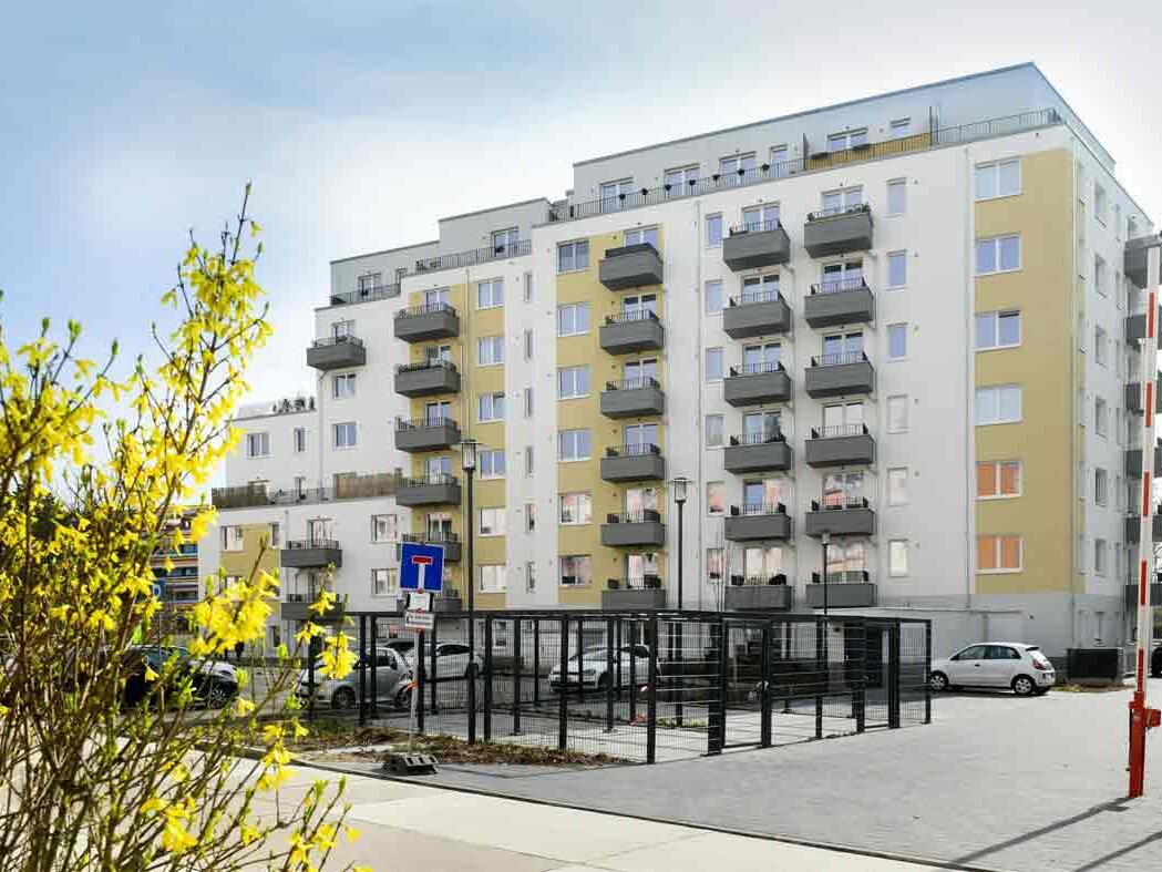 Außenansicht degewo-Neubau Schützenstraße 11 A bis C mit Parkflächen, geöffneter Schranke, Straßenlaterne und blühenden Zweigen links im Vordergrund.